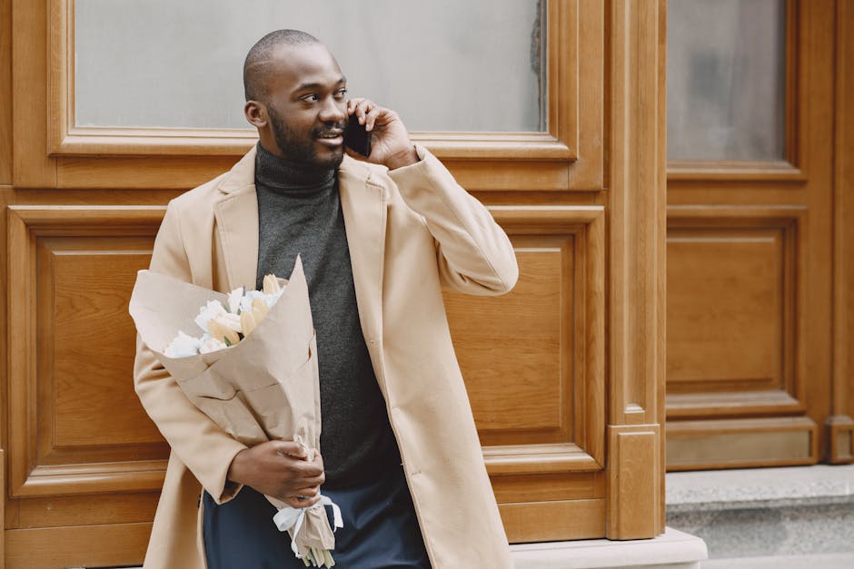 A smiling man with a dark beard and short hair, dressed in a beige coat and dark turtleneck, stands outdoors against a wooden-paneled building. He is holding a bouquet of fresh white tulips wrapped in