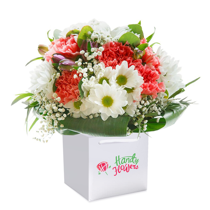 Perfectly composed within a pristine square white gift bag, this radiant bouquet brings together a harmonious mix of colours and textures that echo the vibrancy of Willesden's local parks and high streets. Central to the arrangement, soft coral-pink and peach carnations unfurl with abundant, ruffled petals, their palpable warmth balanced by the elegant purity of crisp white daisies with lively lime-green centres. Interspersed delicately, sprigs of fine baby's breath add a whimsical, airy lightness-much like the dance of dappled sunlight among the trees in Gladstone Park. Deep, glossy green leaves encircle the bouquet, sculpting a natural, protective wrap that grounds the pastel brilliance of the design. Tucked subtly among the blooms, hints of purple-tinged buds can be glimpsed, adding a gentle suggestion of transformation and anticipation. Designed for ease of gifting, the white bag with its sturdy ribbon handles speaks to thoughtful presentation and ease of delivery to homes throughout Willesden, Dollis Hill, Kensal Green, and beyond. Set against a flawless white background, the photo captures every fresh detail in vivid focus, from the tactile texture of carnation petals to the luminescent daisy faces. This bouquet stands as a testament to modern Willesden floristry-ideal for commemorating birthdays, anniversaries, or simply sharing a moment of joy within the vibrant local community.